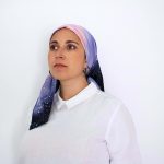 Hand-painted silk headscarf named Cosmic Bloom with cosmic night sky design in purple, orange, and black tones, worn as a Jewish mitpachat