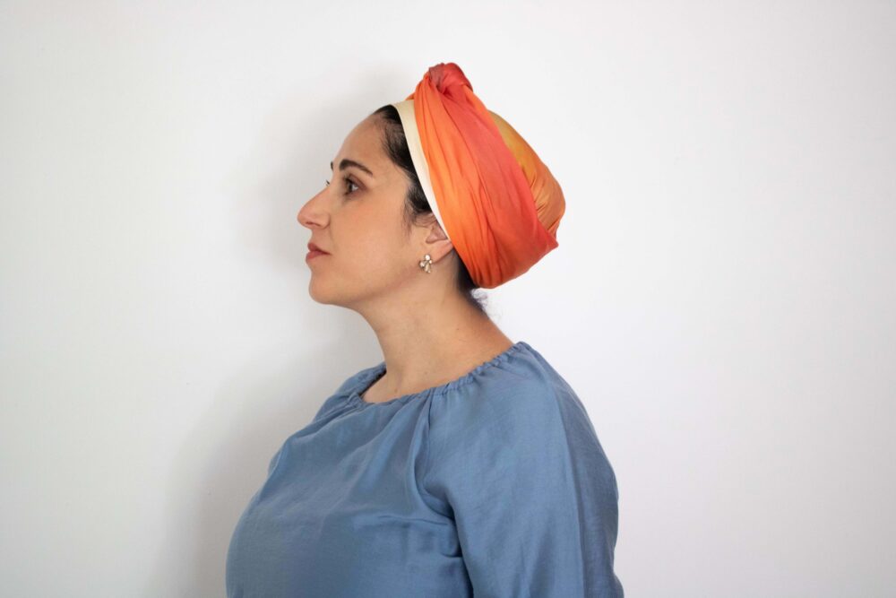 Hand-painted silk headscarf in warm orange and cream tones, displayed hanging vertically titled "Skyfire"